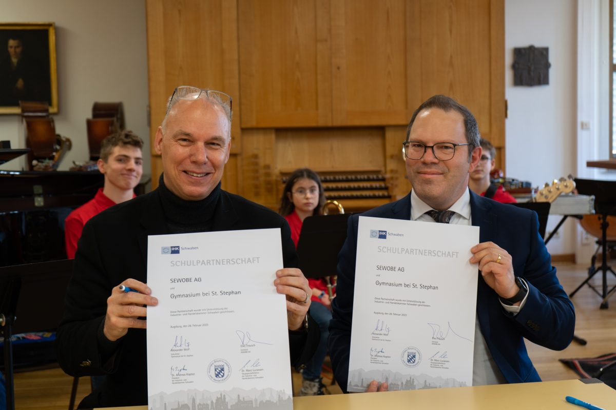 Von rechts Eiko Trausch (Vorstand SEWOBE AG) und Alexander Wolf (Schulleiter Gymnasium bei St. Stephan) halten die Urkunde zur Schulpartnerschaft hoch